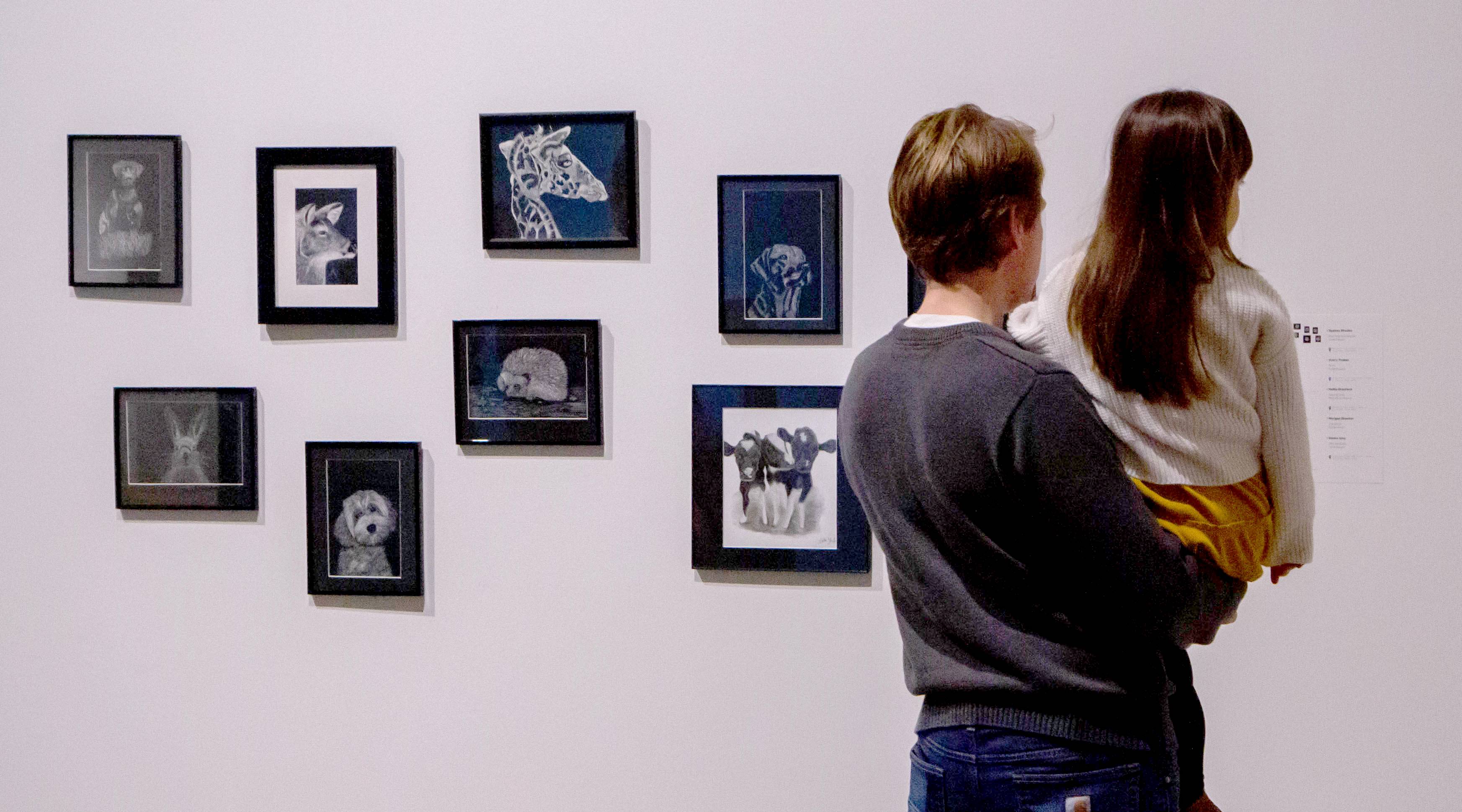 a father holds his daughter up to look at black and white realistic animal drawings