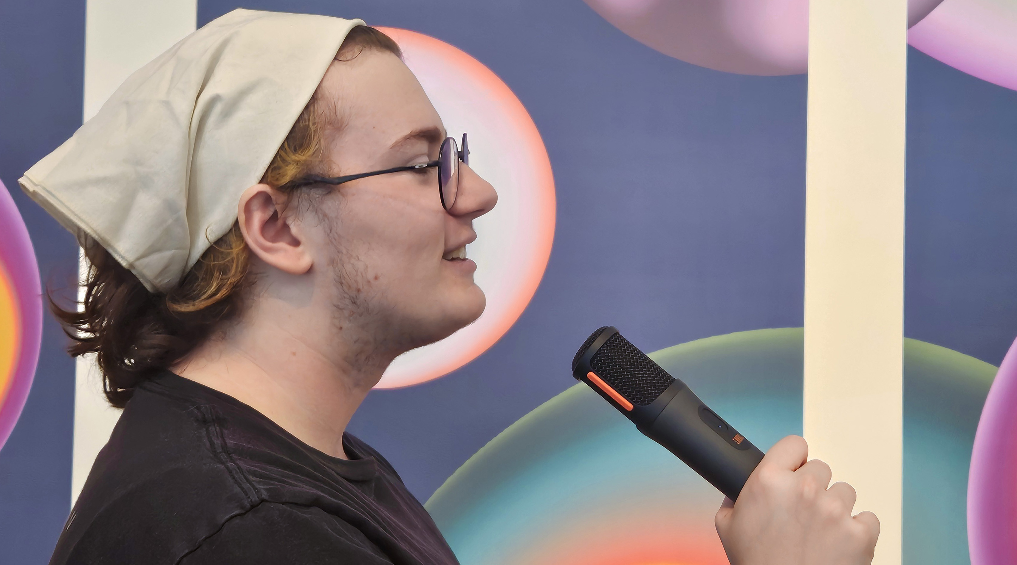 From the side facing right, a man sings with a microphone in front of a blue and purple backgrouns