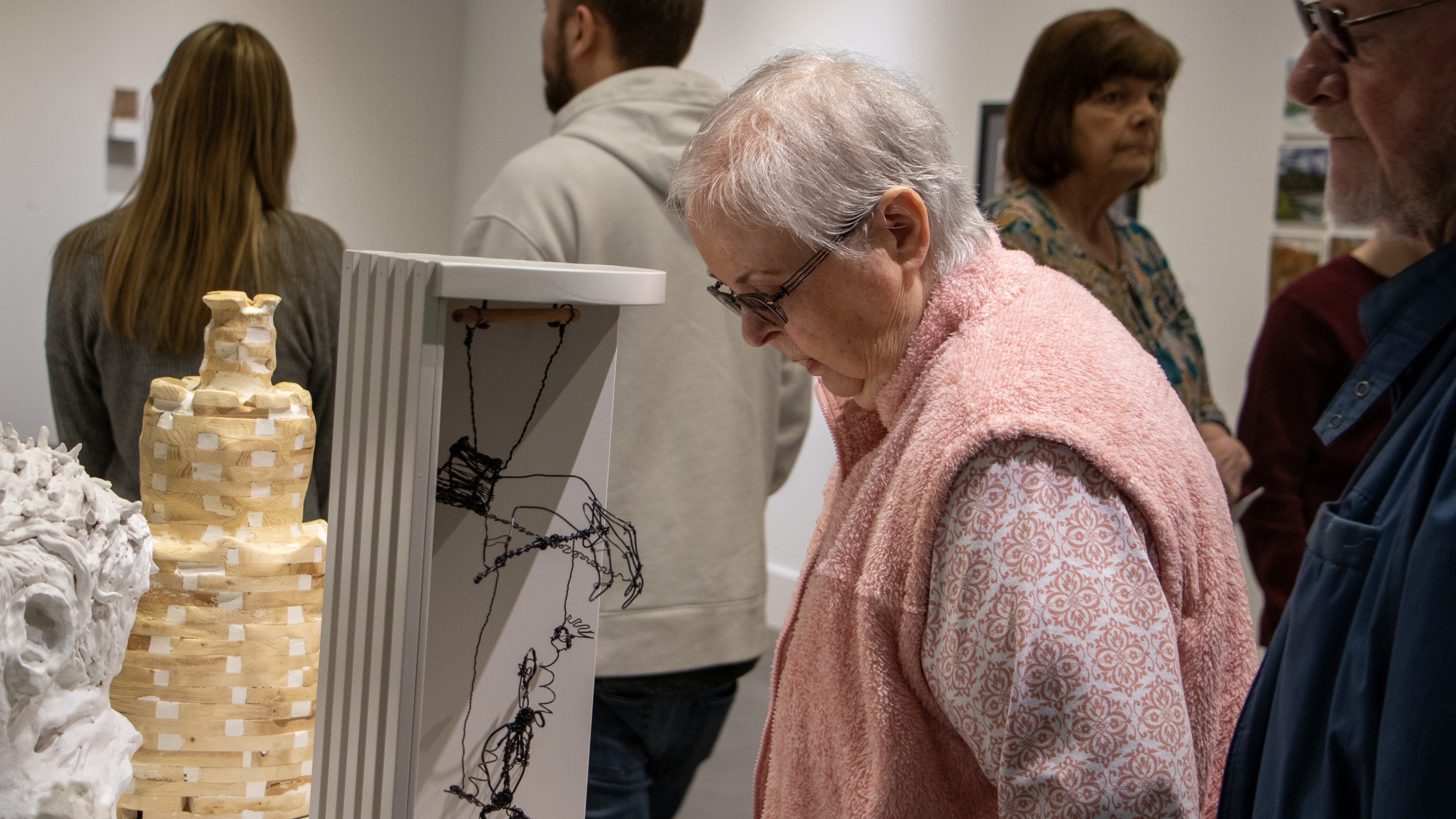 An older woman in a pink sweater looks down at a wire sculpture